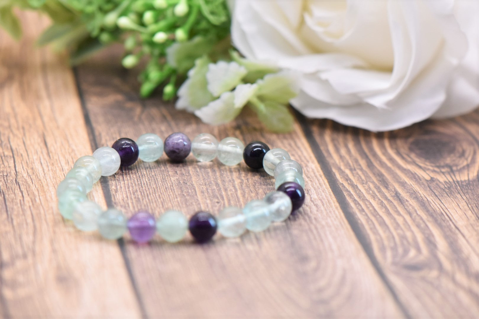 A beaded stretch bracelet made of light green aventurine and dark purple amethyst beads, displayed on a wooden surface with a white rose in the background.