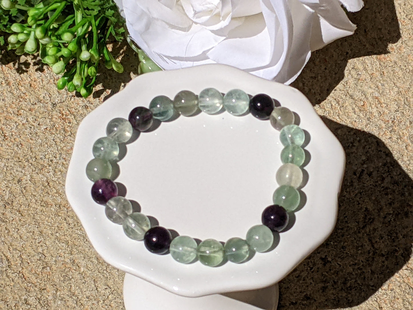 A beaded bracelet made of round translucent light green gemstone beads and a few dark purple beads, displayed on a white ceramic dish over a stone surface with a white flower in the background.